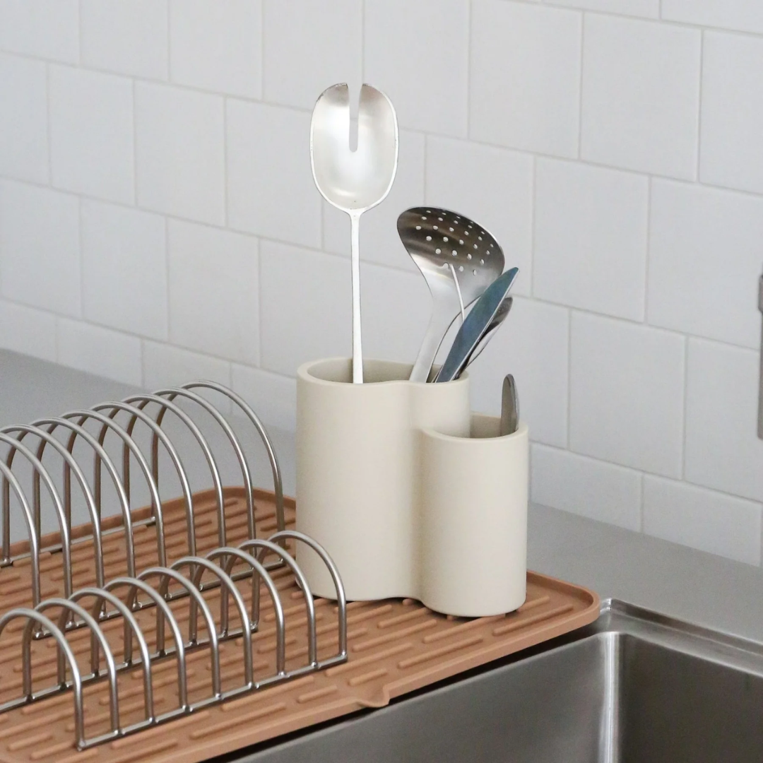 Cremehvid silikone bestikholder med tre rum fyldt med metalredskaber, placeret på en opvaskebakke ved køkkenvasken