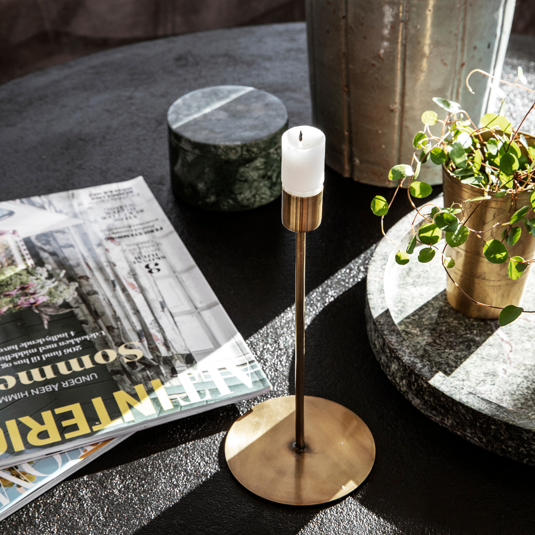 Smal lysestage i antik messing med rund fod og hvidt bloklys, stylet på sort bord med magasiner og planter