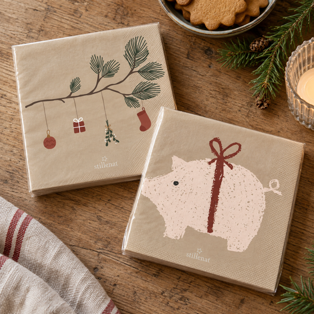 To pakker beige papirservietter med julemotiver: julegris med rødt bånd og grene med pynt