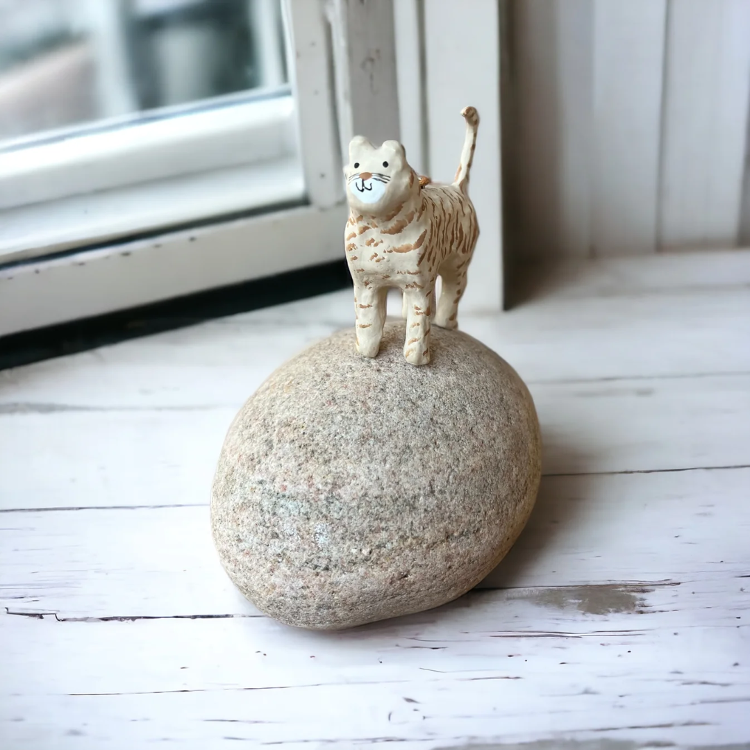 Lille beige papmaché-kattfigur med brune striber, stående på en sten