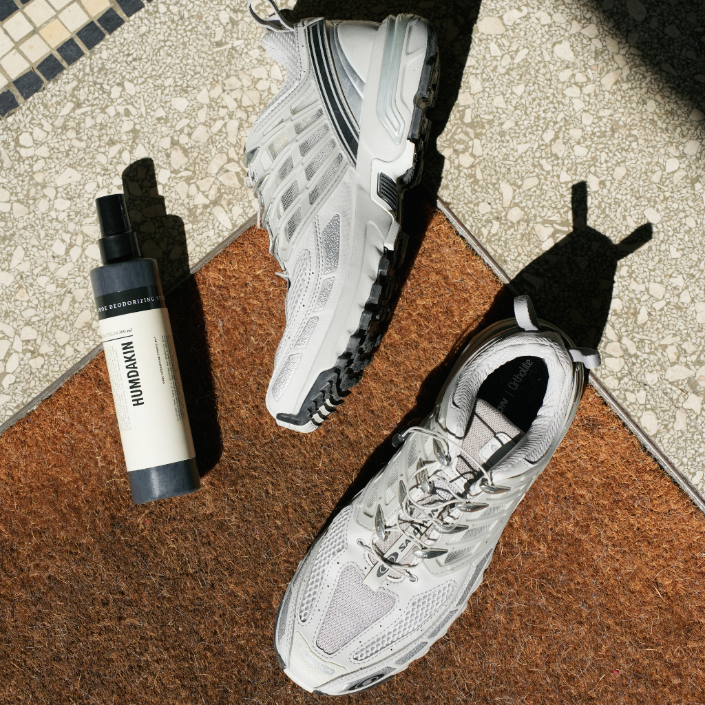 Humdakin skospray i hvid og grå flaske med sort låg, placeret ved siden af et par hvide sneakers