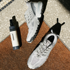 Humdakin skospray i hvid og grå flaske med sort låg, placeret ved siden af et par hvide sneakers