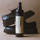 Humdakin skospray 2-i-1 i mørkebrun sprayflaske på 300 ml med beige etiket, foran sorte lædersko