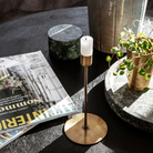 Smal lysestage i antik messing med rund fod og hvidt bloklys, stylet på sort bord med magasiner og planter