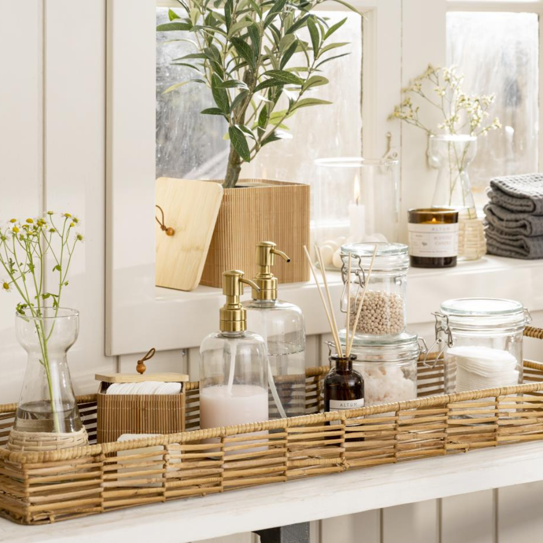 Klar glassæbedispenser med messingfarvet pumpe på rattan-bakke med badeværelsestilbehør