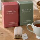 To Supertea æsker i rosa og grøn med et teabrev og en hvid kop te foran